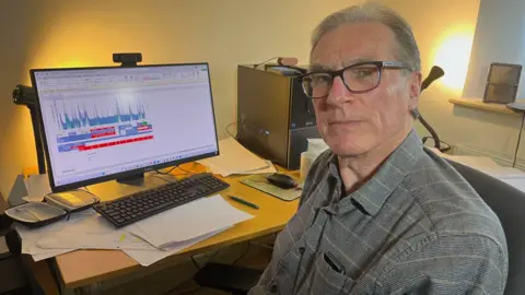 A man with short gray hair and glasses, wearing a gray patterned shirt, sitting at a desk. He looks directly at the camera with a neutral expression. In the background, a computer monitor displays a graph with red and blue lines, along with a keyboard, mouse, papers, and a computer tower. 