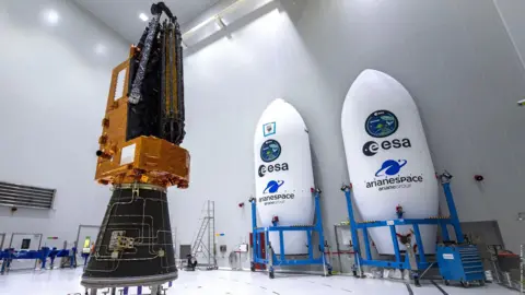 ESA-CNES-ARIANESPACE The biomass satellite sits inside an all-white warehouse. It has a black cone-like base, an orange l-shaped top that has the look of foil, with the antenna attached and furled. To the left of the satellite sit two shells which will hold the satellite, they are white elongated cones with "ESA" written on them, and held in blue scaffolding trolleys.