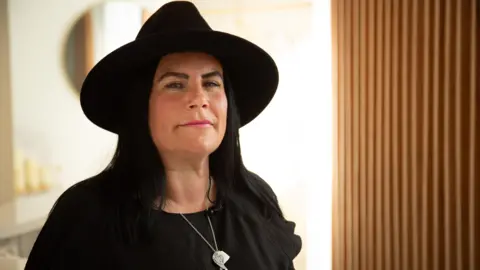 BBC A woman wearing a black hat and a black top, standing near a window and looking at the camera